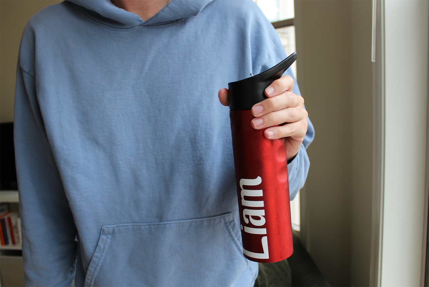 A person in a blue sweatshirt, head not pitched, holds a read metal bottle which reads "Liam."